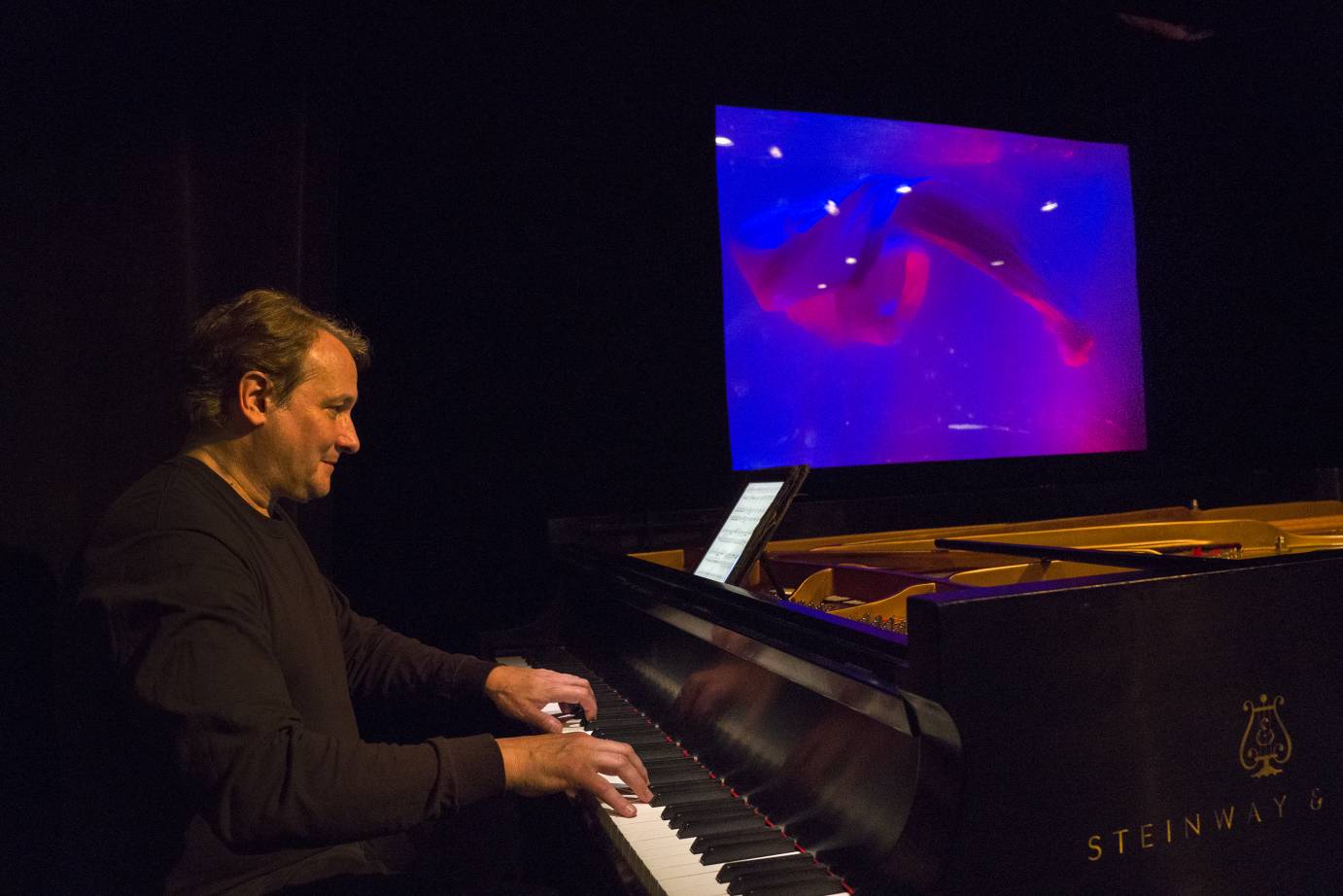 Christopher O'Reilly sits at a Steinway piano in front of a magenta-illuminated aquarium. 